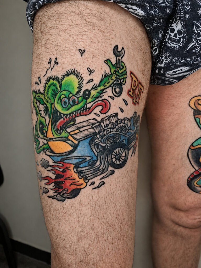 Tatouage Rat Fink old school coloré sur la cuisse, représentant le célèbre personnage Rat Fink en train de piloter une voiture hot rod avec un moteur apparent et des flammes, réalisé par Vittoria Tattoo en Creuse.