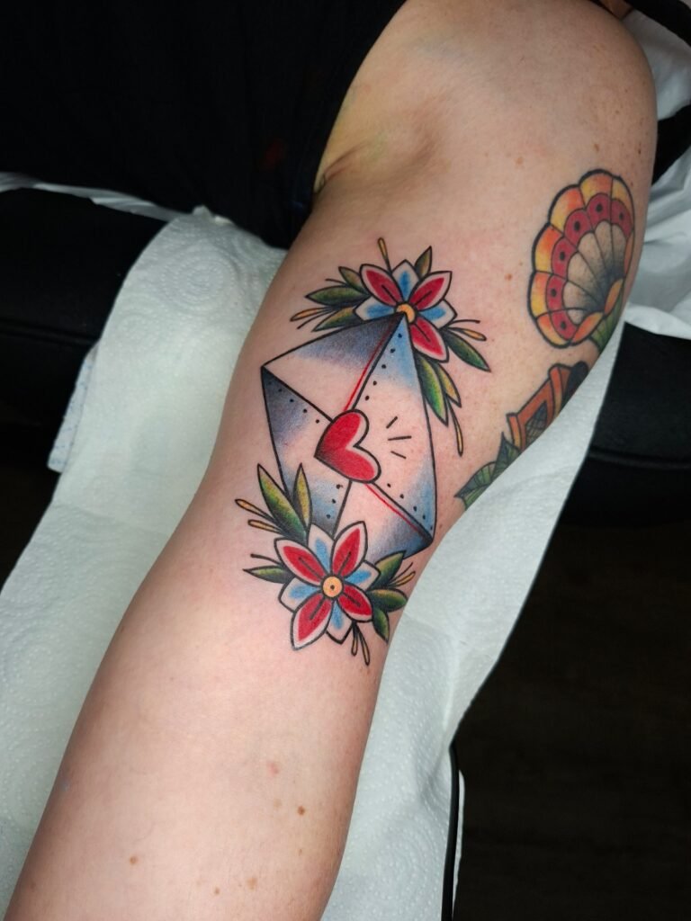 Tatouage old school représentant une enveloppe avec un cœur rouge, entourée de fleurs et feuillages colorés, réalisé sur le bras par Vittoria Tattoo en Creuse.