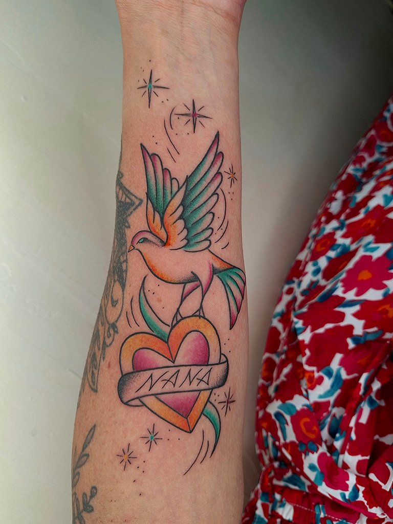 Tatouage Old School représentant une colombe colorée en vol au-dessus d’un cœur avec un ruban portant l’inscription « NANA », réalisé sur l’avant-bras avec des étoiles décoratives.