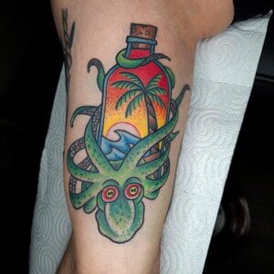 Tatouage old school poulpe vert entourant une bouteille avec paysage tropical (palmier, vague et coucher de soleil) sur mollet – réalisé chez Vittoria Tattoo.