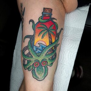 Tatouage old school poulpe vert entourant une bouteille avec paysage tropical (palmier, vague et coucher de soleil) sur mollet – réalisé chez Vittoria Tattoo.